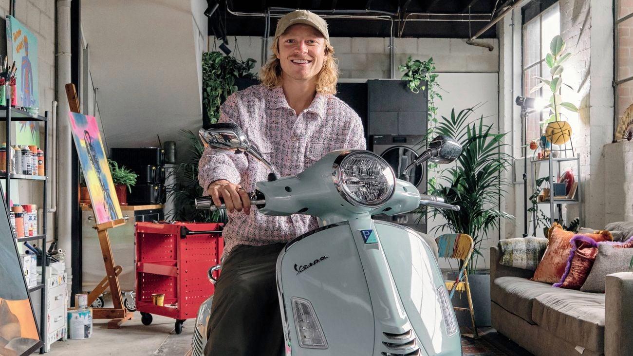 A man with long blonde hair and a cap sitting on a light blue scooter in an art studio