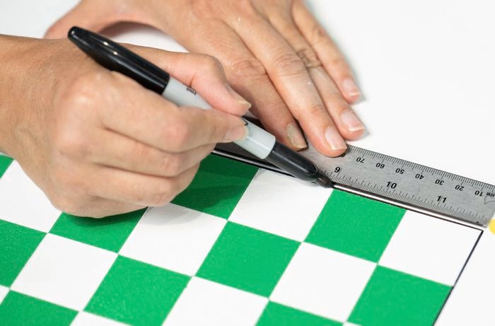 A person is drawing a neat border around the painted green and white snakes and ladders grid on the toy box lid using a ruler and marker. 