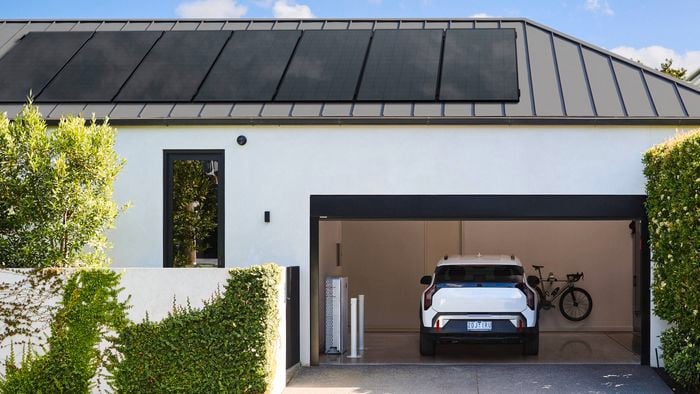 Modern residential home exterior with a low-pitched metal roof fitted with solar panels, featuring an open double garage with a white SUV parked inside, a bicycle mounted on the back wall, and neatly landscaped greenery along a concrete driveway.