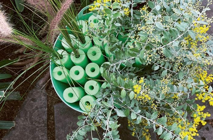 Hands filling a green tub, containing green pool-noodle pieces, with various plants and grasses.