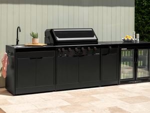 A black outdoor kitchen with a barbecue, sink and bar fridge in it, sitting on paving outside.