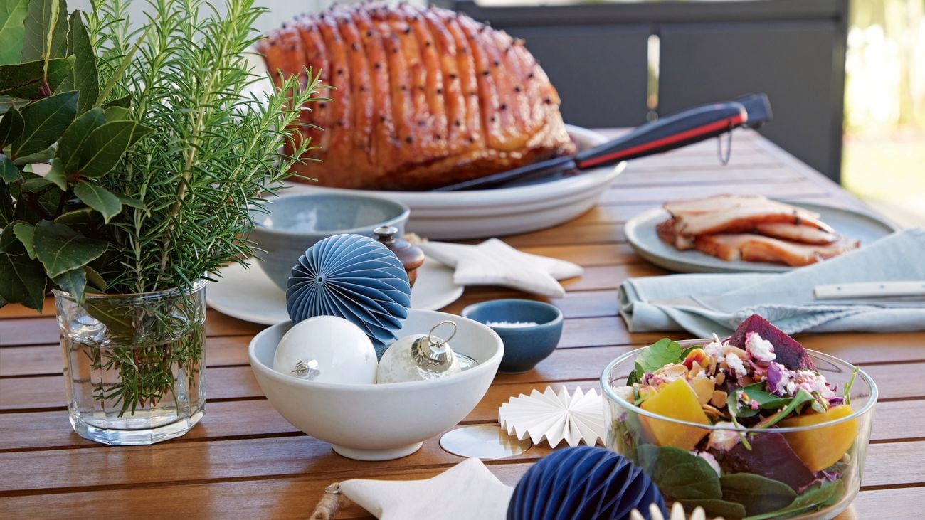 An outdoor table decorated with blue and white, paper baubles sit in front of a Christmas ham.