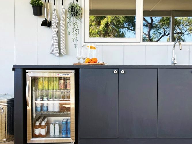 "Outdoor kitchen with black countertop and cabinets, featuring a small fridge stocked with drinks, a wall-mounted light, hanging plant, kitchen towels, and a window overlooking trees above the sink