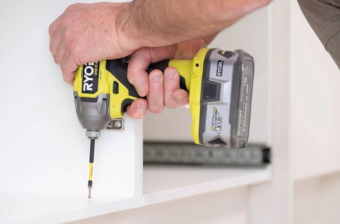 A person is using a power drill to pre-drill through the cabinet base and into the kickboard to prevent splitting.    