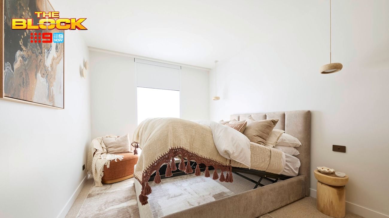 A furnished bedroom with a bed tilted above the frame, carpet, bedside table, and chair included in it and The Block logo included on the image.