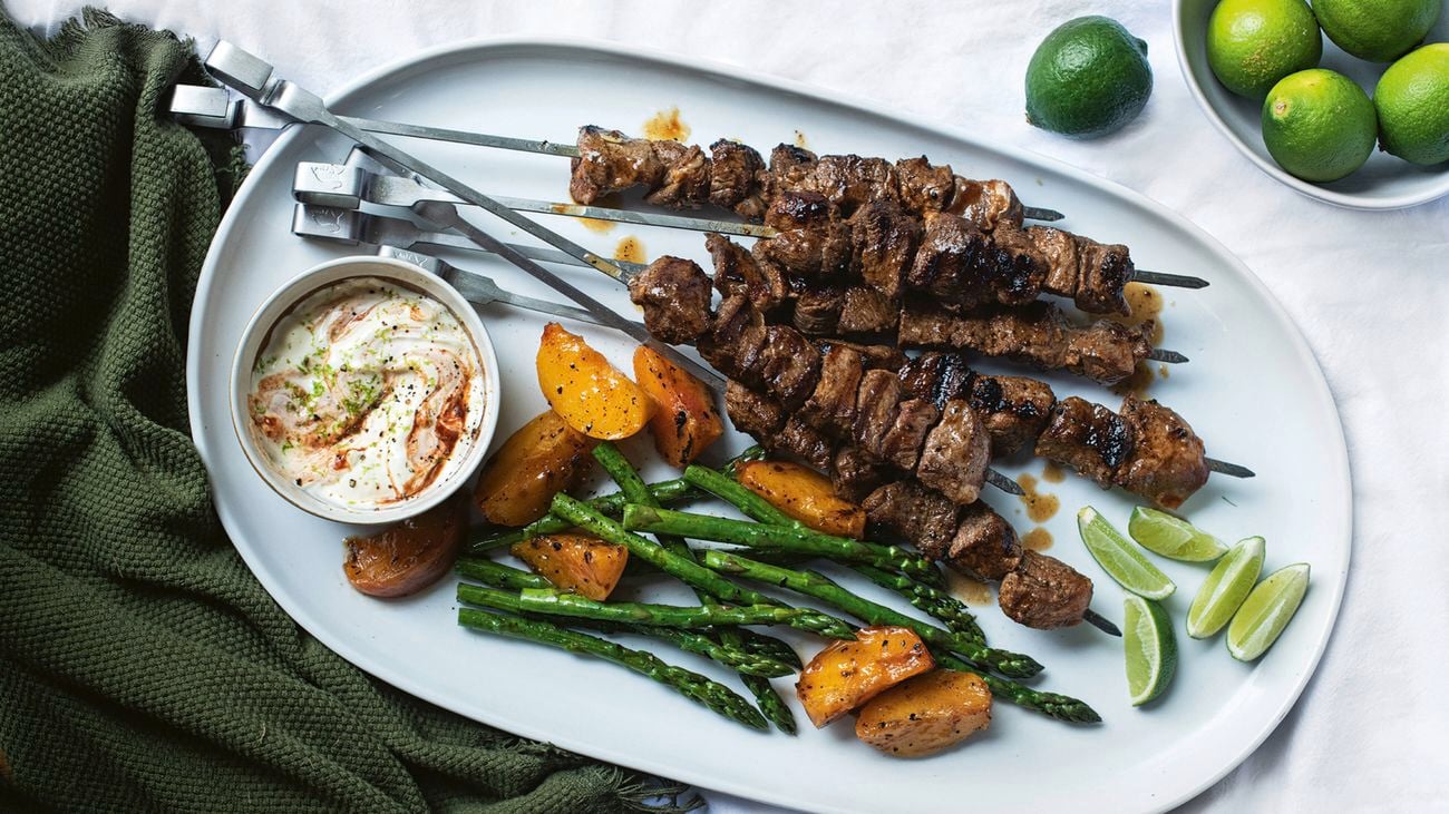 Smoky lamb and peach kebabs with asparagus spears, lime wedges, and a small bowl of yoghurt sauce on a white oval plate in a white tablecloth with green napkins.