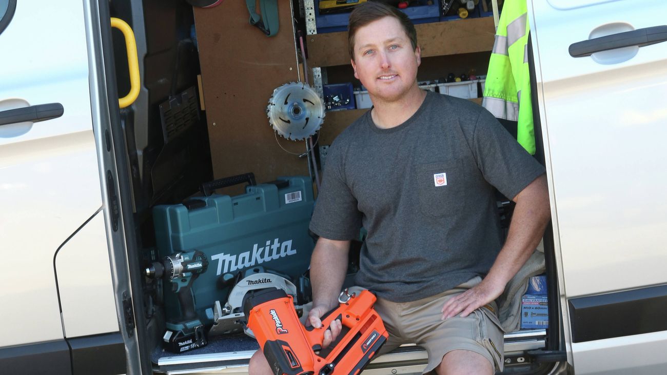 A tradesperson in a grey Citeco t-shirt and khaki shorts, holding a Paslode nail framer sits in the back of a van onsite.