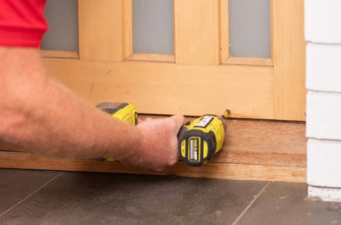 A hand is drilling holes at the bottom of the door to install screws.