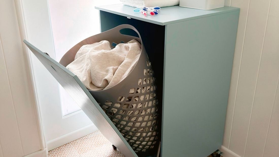 A light blue tilt-door laundry hamper cabinet with a grey hamper inside, holding white laundry.