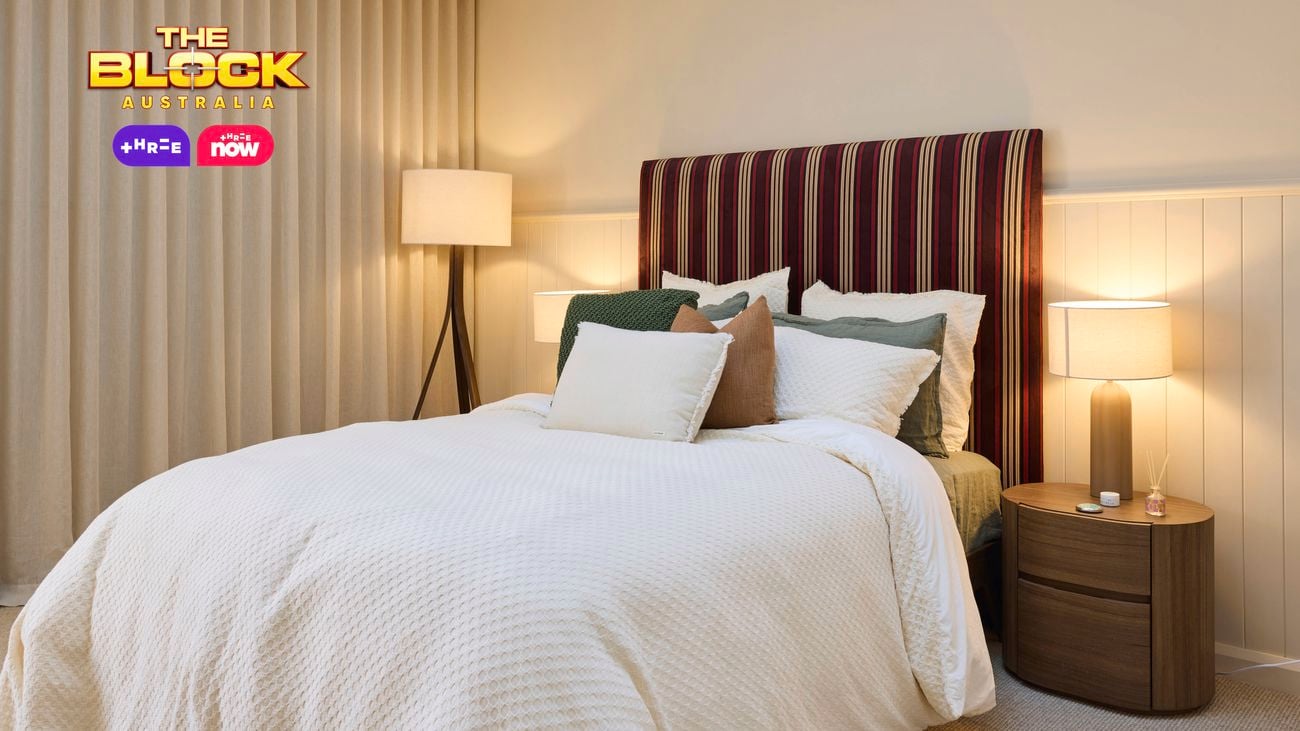Cream bedroom with a double bed and a patterned bedhead, white linens and a lamp in the corner and a wardrobe opposite.