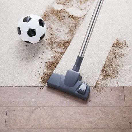 A vacuum cleaner creating a clean patch through a wide patch of loose soil around a soccer ball over carpet and wood flooring