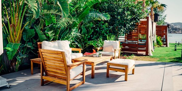 Outdoor patio with wooden lounge furniture and white cushions, surrounded by lush tropical plants and palm trees in a landscaped garden.