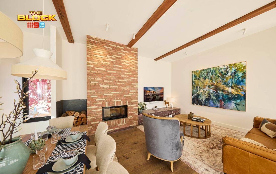 A modern living room featuring a central brick fireplace, exposed wooden ceiling beams, and a large abstract painting on the right wall. Furnished with a brown leather sofa, two gray armchairs, and a round wooden coffee table atop an area rug. A dining table set for a meal is visible to the left. 