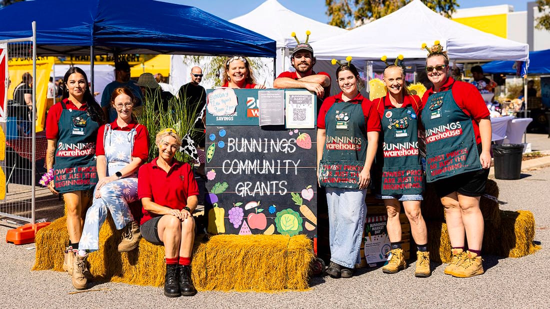 Group of Bunnings Team Members standing and sitting around a Bunnings Community Grants sign at an outdoor community event with market tents in the background.