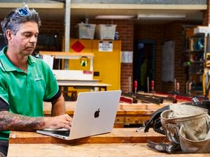 A tradesperson in a green and black polo shirt sits at a wooden workbench using a silver laptop, with tools and workshop equipment visible in the background.]