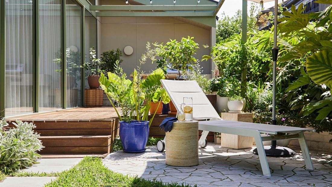 An outdoor patio with a white sunlounge next to a small side table holding a jug and a glass. A wooden deck and sliding glass doors sit on the left with a dense garden and lush foliage on the right.