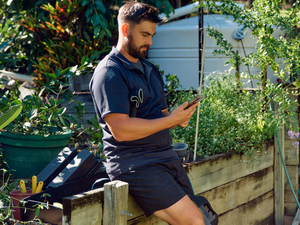 Alt tag: A tradie A tradie wearing a dark shirt and shorts sits on a wooden garden bed looking at his mobile phone, surrounded by plants and gardening tools
