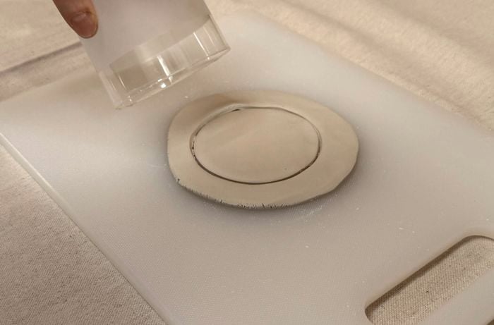 A hand pressing a cup into a flat piece of clay to form a circular outline on a white surface.