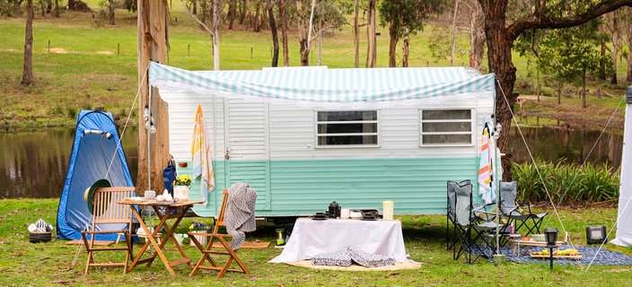 White and light blue caravan in rural grassland alongside a river surrounded by camping products including a table and chairs, camping chairs, tent and picnic rug 