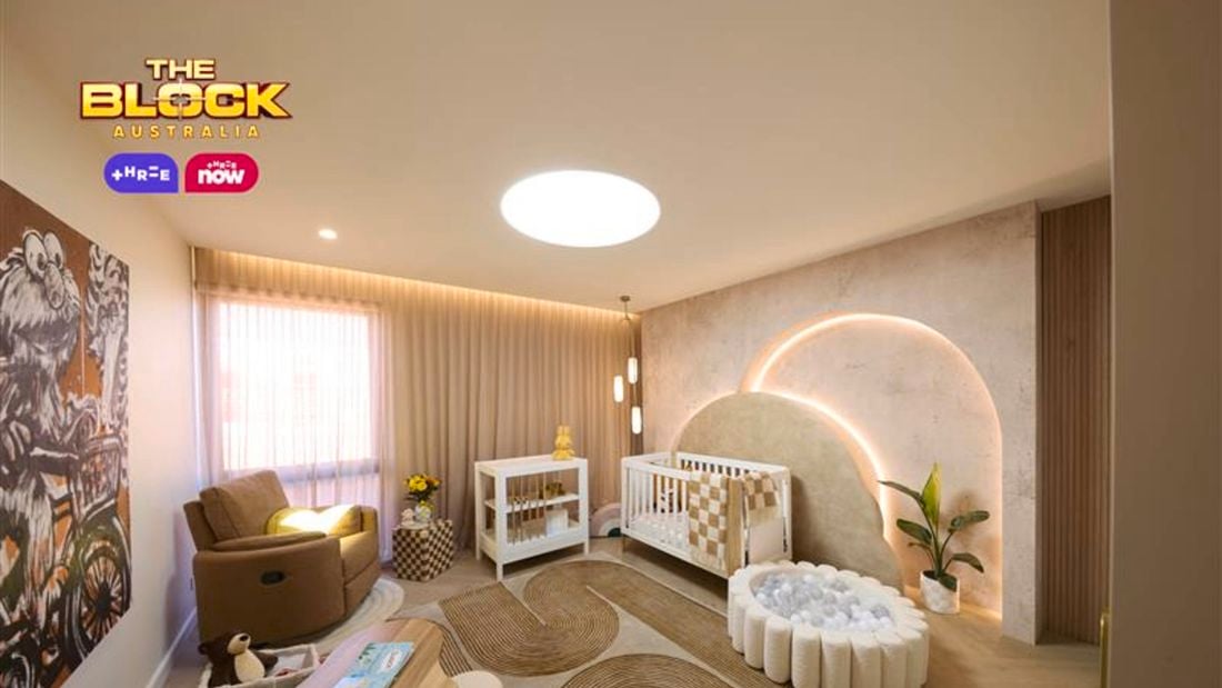 Neutral baby bedroom with brown and tan colours. Showcasing a cot and change table in front of an arched feature wall with feature lighting. There is a large chair in the corner and a play area on an arched pattern rug.