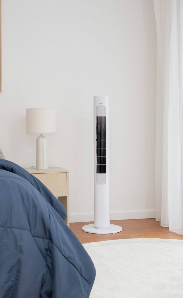 White tower fan standing beside a bed in a bright bedroom with sheer curtains, a bedside table, and a framed wall print