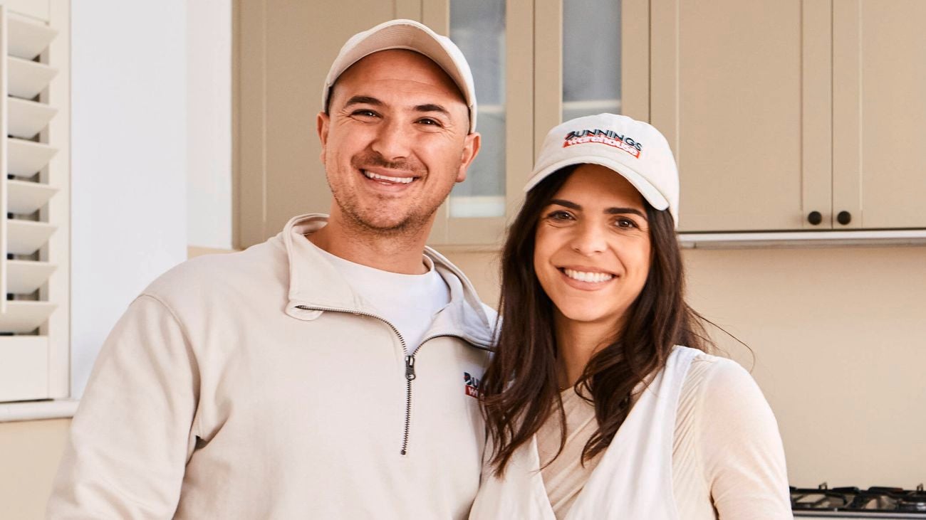   A couple consists of a man and a woman standing side by side behind a kitchen island in a modern beige kitchen, both wearing branded caps and casual tops, with cabinets, a cooktop, coffee machine, and stainless steel fridge visible in the background.