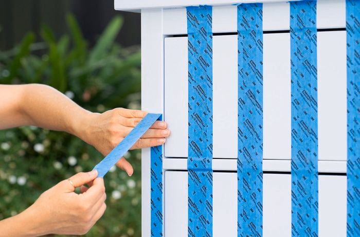 Hands applying blue painter’s tape in vertical strips across the front of a cabinet drawer.