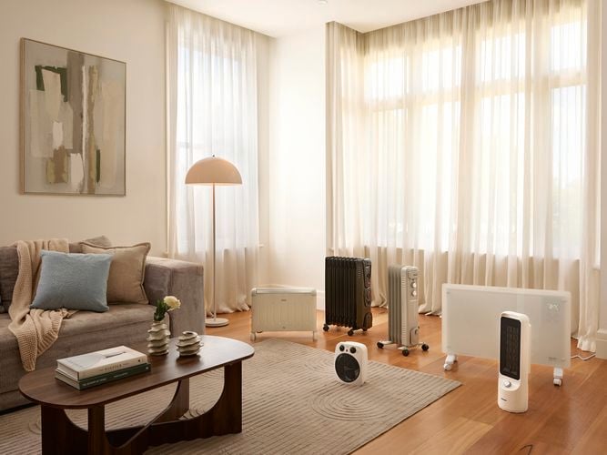 A furnished lounge room with many heaters in it, a painting on the wall, curtains in front of the windows with timber flooring.