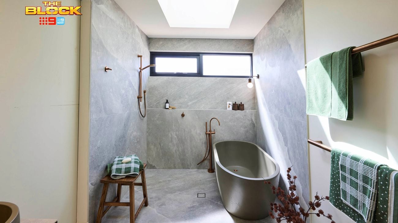 Modern bathroom with freestanding tub, walk-in shower, grey stone tiles, skylight, copper towel bars, and wooden stool with towels. 