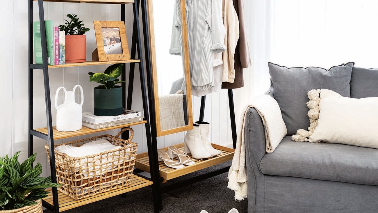 A couch, stand with shelves on it and a stand with a mirror and a hanging rail on it with clothes hanging from it