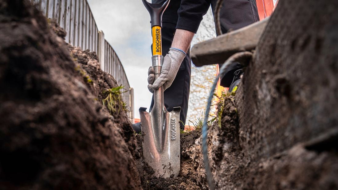 A close-up, ground-level view of a person using a metal spade to dig a trench outdoors, with soil on either side and part of a wheelbarrow visible in the foreground.