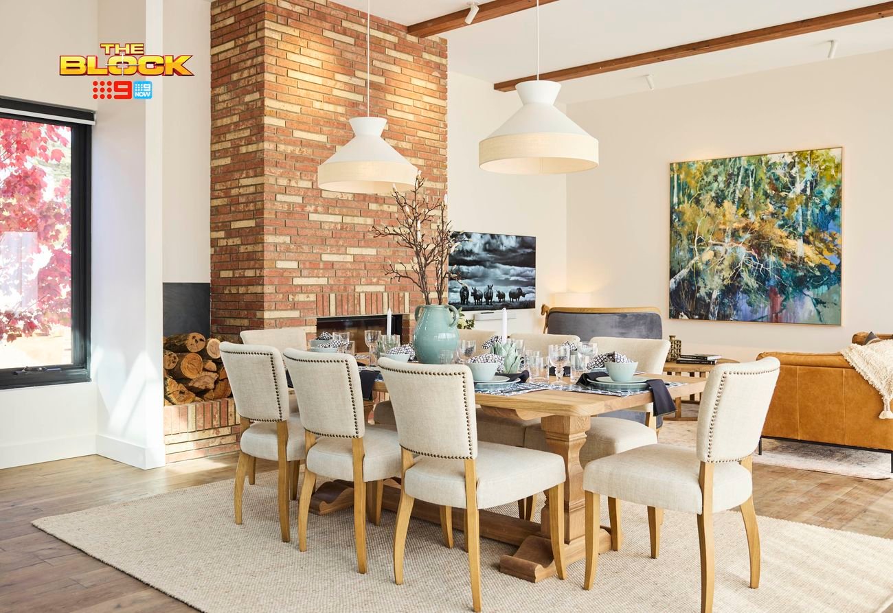 A stylish dining and living room featuring a wooden dining table set with plates, glasses, and a centerpiece, surrounded by six upholstered chairs. A brick fireplace with stacked logs sits nearby, with two pendant lights overhead. A brown leather sofa with cushions and a throw blanket faces a large colorful painting. A window reveals red foliage outside. 