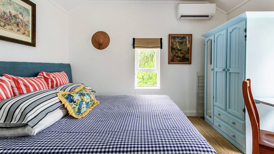 Bright bedroom with a blue upholstered bed, check-pattern bedding, light blue wardrobe, workstation and garden-view window beneath a vaulted white ceiling.