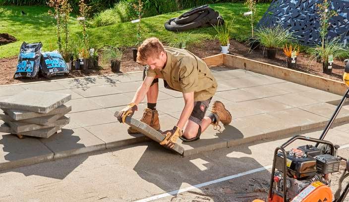 A landscaper laying pavers in a backyard with lawn and plants.
