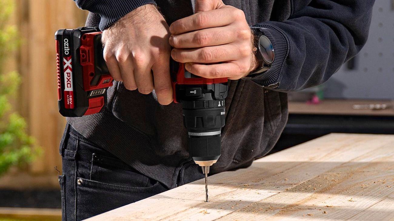 Close up of hands holding a cordless drill as a screw is driven into a piece of timber, with loose screws nearby