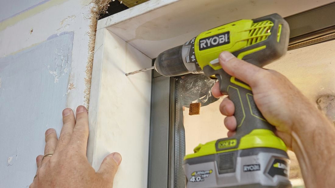 A person drilling through the side of a window frame fitted into a plaster wall