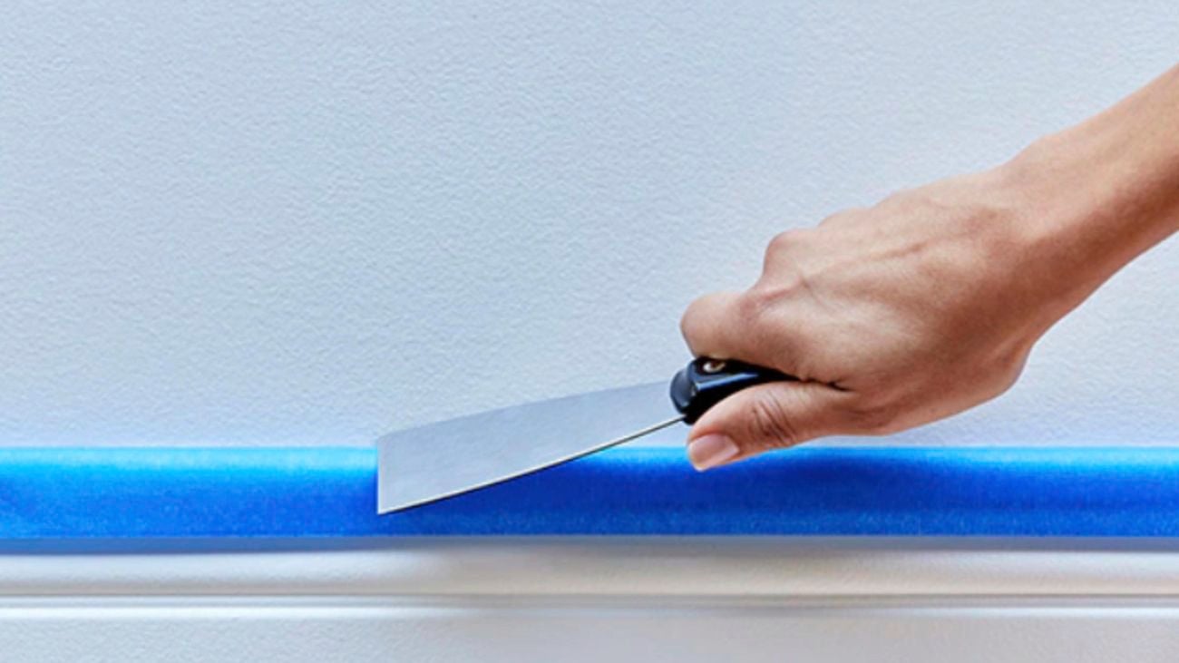 A hand using a narrow scraper to press blue Scotch tape to the top edge of a skirting board