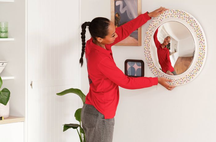 A Bunnings Team Member is carefully hanging a round decorative mirror on a wall-mounted picture hanger, checking its position to make sure it sits straight and secure. 