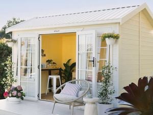 An angled view of a large timber shed painted white with a front deck featuring hanging baskets, pot plants and a chair sits in a garden setting. The open French doors show the yellow interior, a desk and stool.