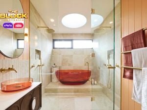 Modern bathroom with amber tub in glass shower, beige tiles, dual overhead showerheads, amber basin with round mirror, and gold towel rack. 
