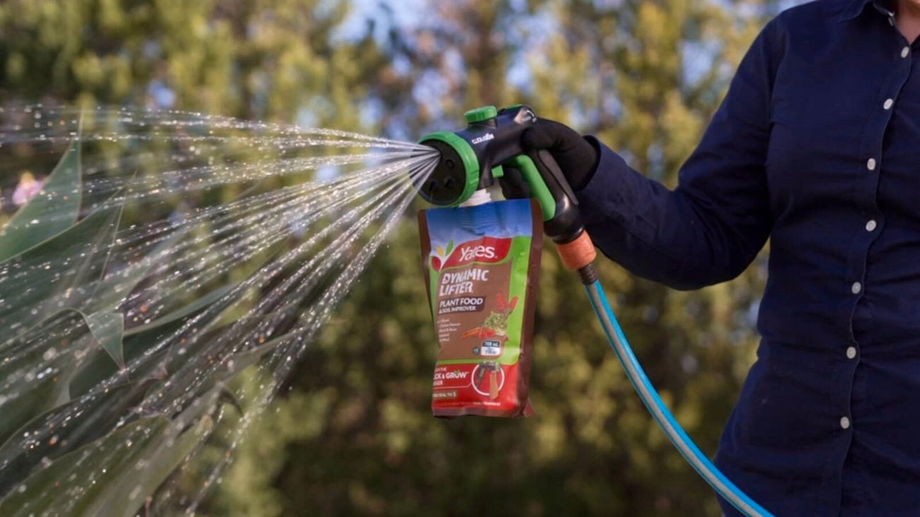 Person holding a hose spray gun with a Yates Dynamic Lifter plant food pouch attached, spraying water onto garden plants.