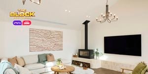 A modern living room with high ceilings, featuring two chandeliers, a wall-mounted flat-screen TV, and a fireplace with logs stored below. The space includes a light-colored sectional sofa with cushions and throws, two armchairs with green cushions, and two round wooden coffee tables. Abstract artwork hangs above the sofa, and large windows bring in natural light. Decorative plants are placed throughout. 