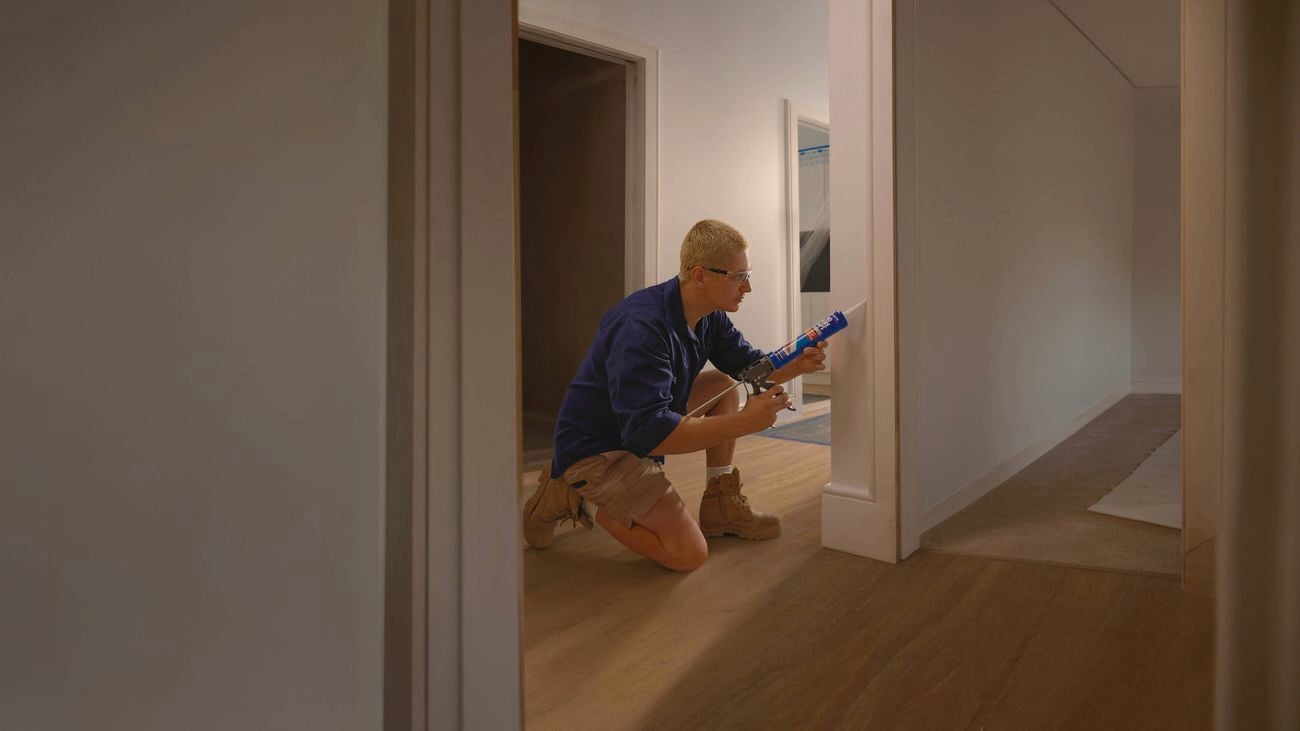 A tradesperson in a navy blue shirt, khaki shorts and wheat coloured boots uses Selley’s No More Gaps on edging in a hallway of a house.