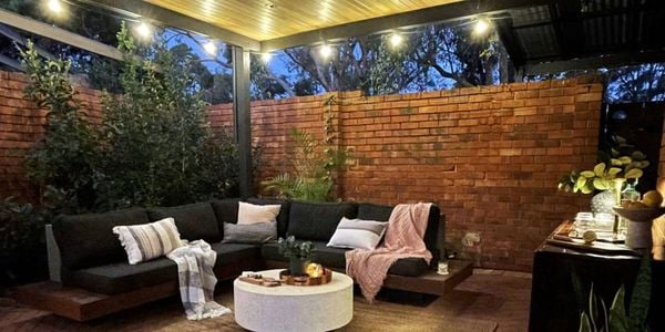 Outdoor timber floating lounge with grey cushions, throw rug, coffee table sitting in front with red brick wall in back ground