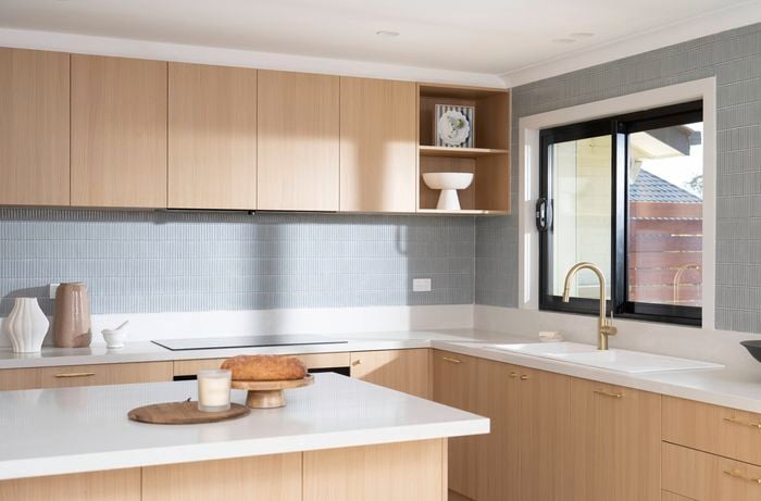 A bright kitchen with natural sunlight features wood cabinets, a gold faucet, a large window, and a clean white countertop with decorative items.  