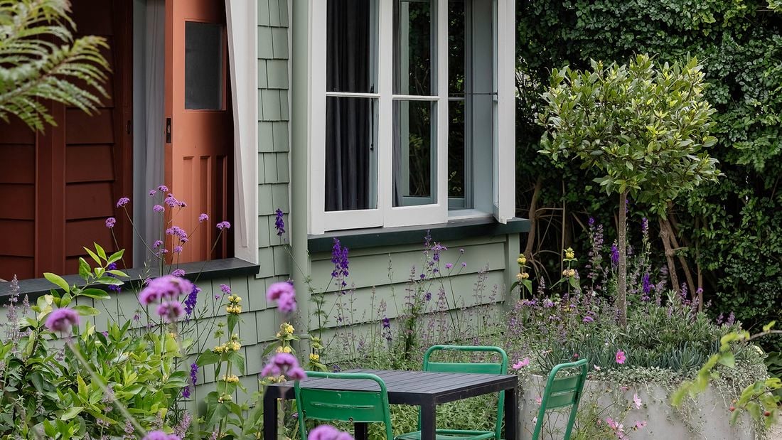 A weatherboard house painted with Dulux paint colours pale green with white trims. Small courtyard with a black table and four green chairs and purple flowers.