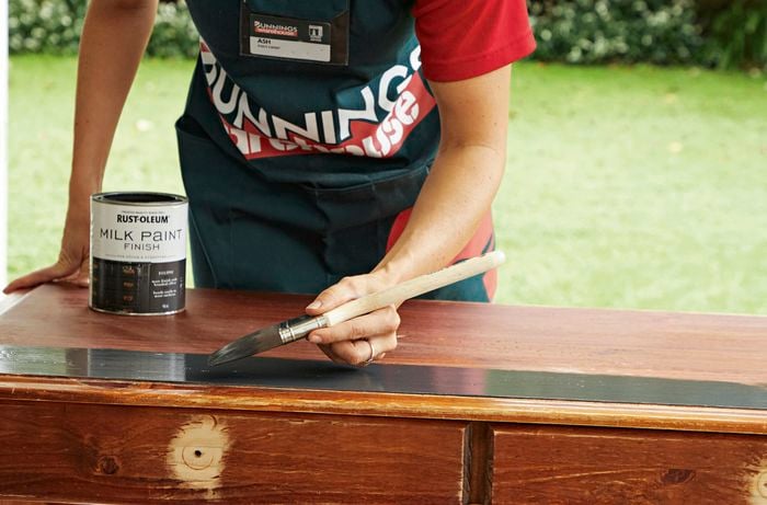  A woman brushes Rust-Oleum milk paint onto a timber desk using a paintbrush.
