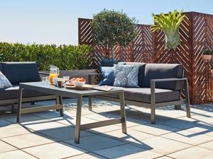 A grey stained coffee table with two matching outdoor lounges in a paved outdoor area, with a breakfast tray on the table with a pitcher of orange juice