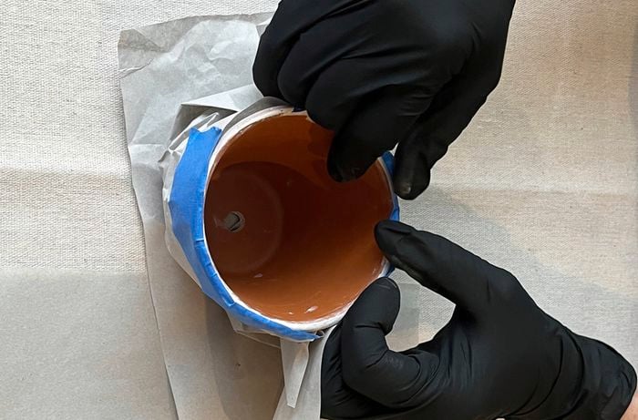  A pair of hands wearing black gloves covers painted pots with paper using painter’s tape, on a drop sheet.