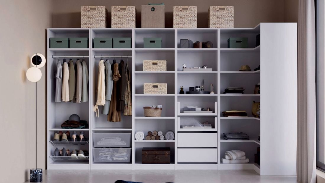 A large open wardrobe unit with shelves and hanging rails holding clothes, shoes, folded towels, storage boxes, wicker baskets, drawers and boxes on top. A floor lamp stands on the left.